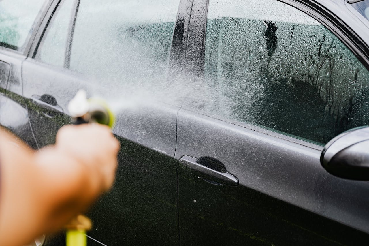 A car being washed with a hose outdoors. Water sprays create a cleaning effect on the vehicle.